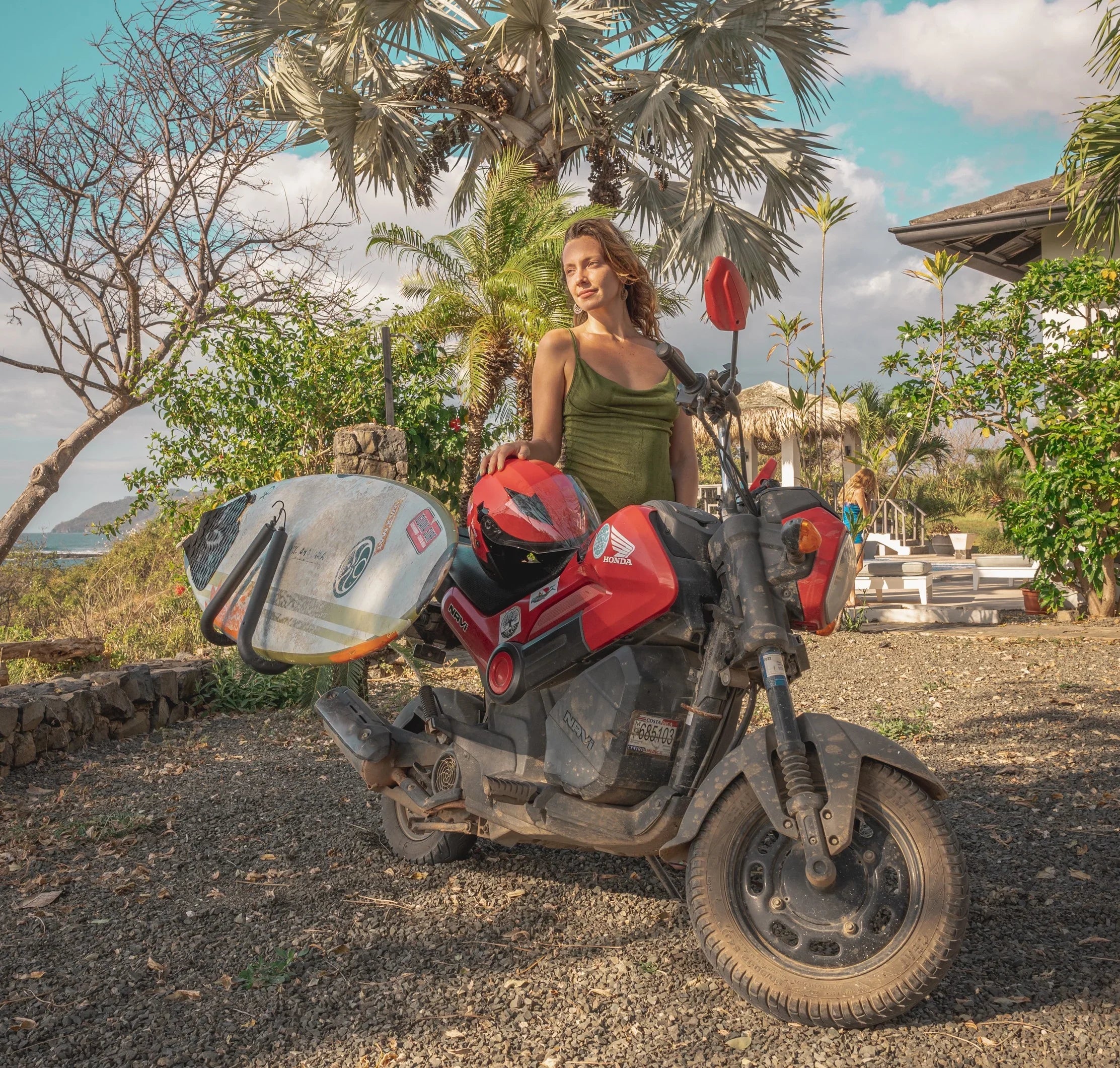 Woman with red Honda scooter and surfboard in Tamarindo, Costa Rica, near palm trees and ocean view